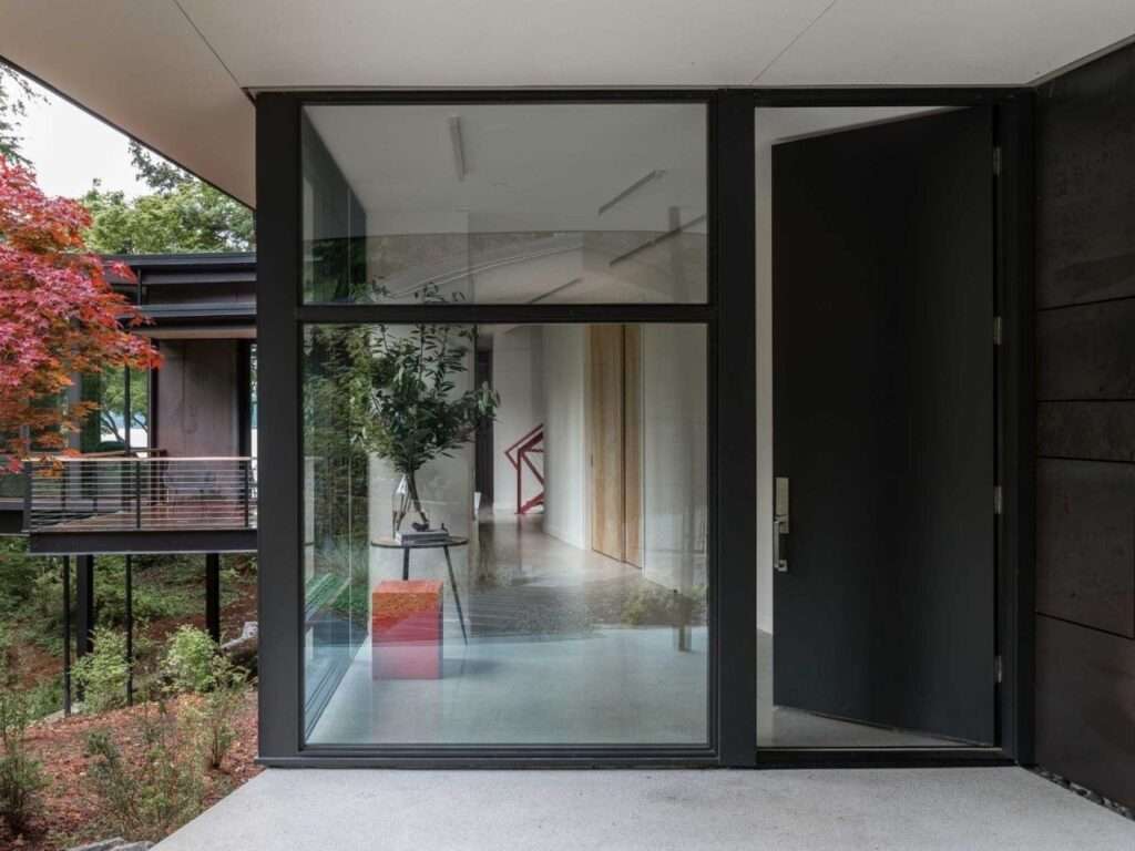 Modern residential entrance featuring a large black pivot door, floor-to-ceiling glass paneling, and a glimpse of the red staircase inside.