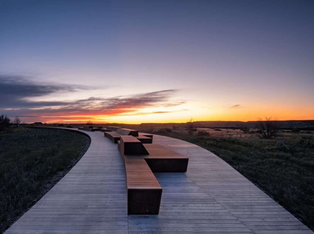 A wooden outdoor boardwalk curving through the prairie landscape at sunset with modern seating benches.