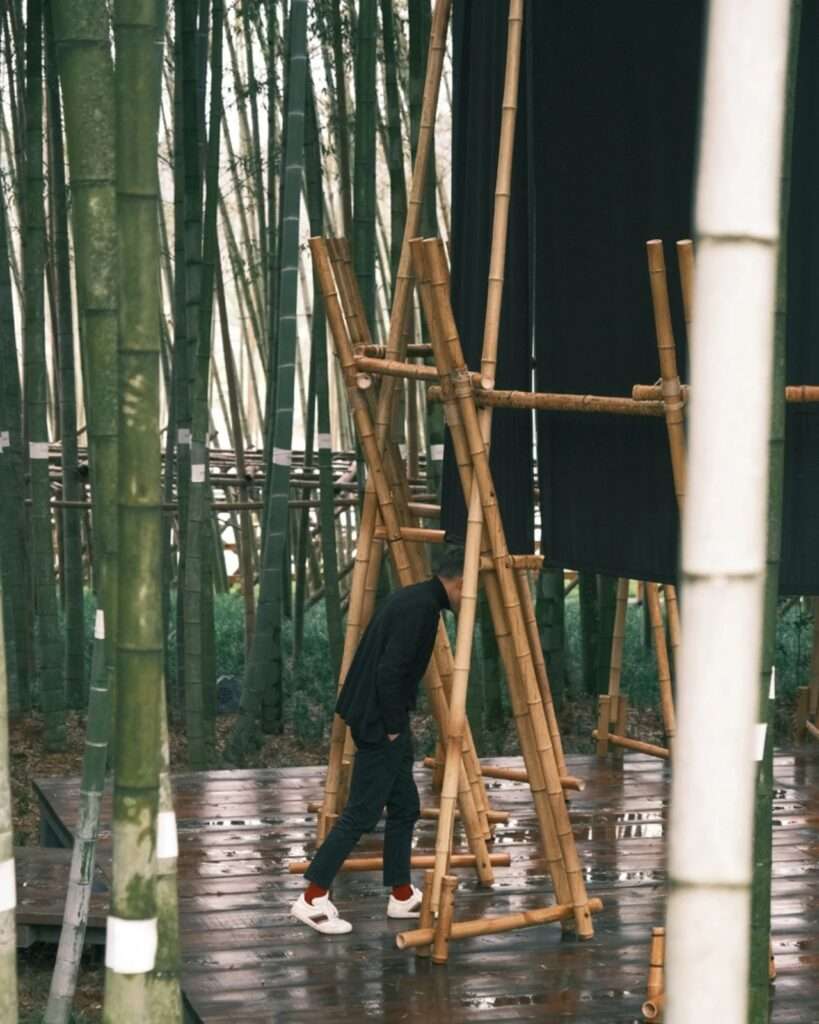 A visitor bending forward to walk underneath a low-hanging horizontal bamboo beam to enter the pavilion on a wooden deck.