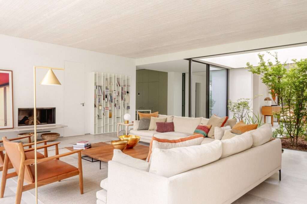 An open-plan interior featuring a large white sectional sofa, a fireplace, leather lounge chairs, and a white bookshelf, organized around a central glass-enclosed courtyard with trees.