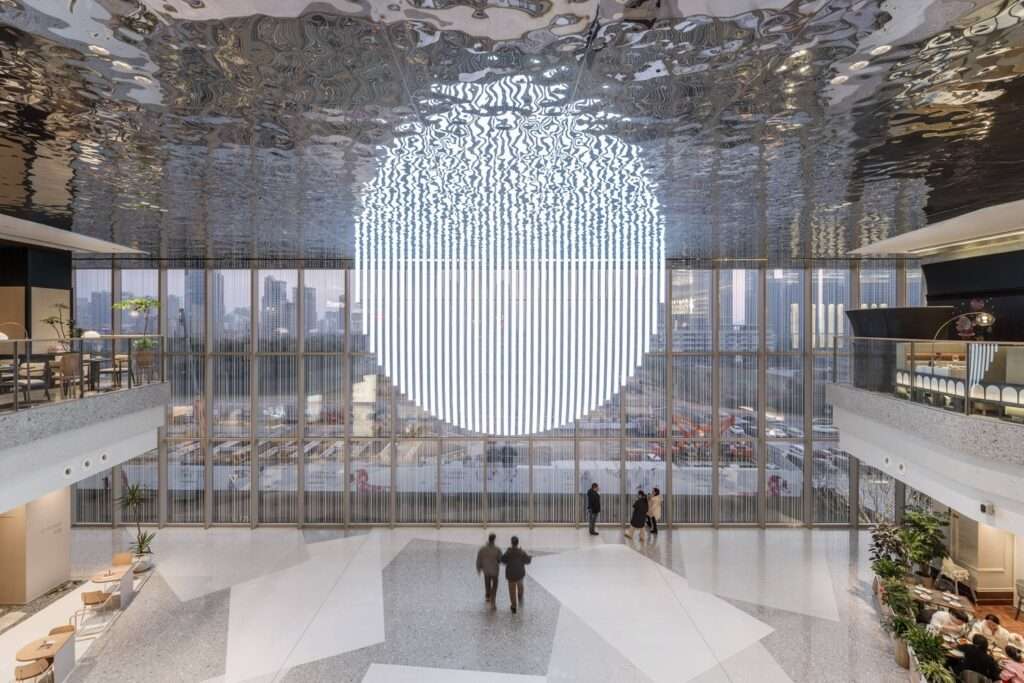 The Moon Garden event space at Wuxi MixC Mall featuring a semi-circular light sculpture reflected on a rippled stainless steel ceiling next to a large glass curtain wall.