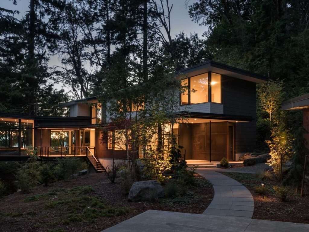 Two-story modern house illuminated at twilight, nestled into a dense hillside forest with a stone pathway leading to the entrance.