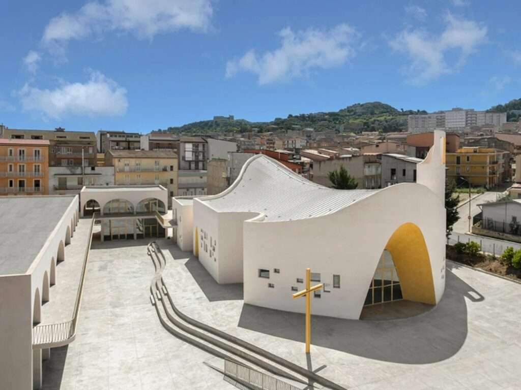 High-angle shot of the Santa Barbara Parish Complex showing the undulating metal roof, central courtyard, and the town of Licata in the background.