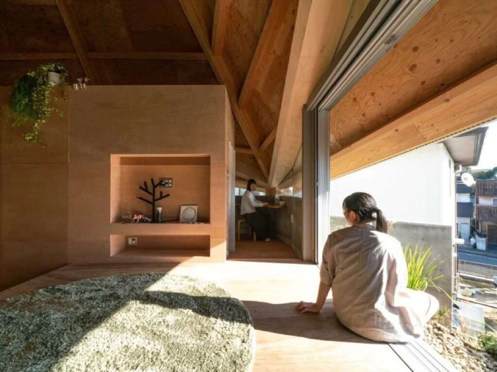 Interior view looking out toward a sunny terrace where a resident sits on the wooden floor ledge next to large open windows.