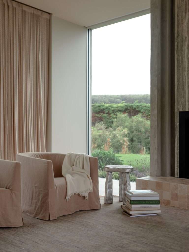 Soft-toned fabric armchairs and textured drapes inside a cozy living space facing a floor-to-ceiling window frame with coastal views.