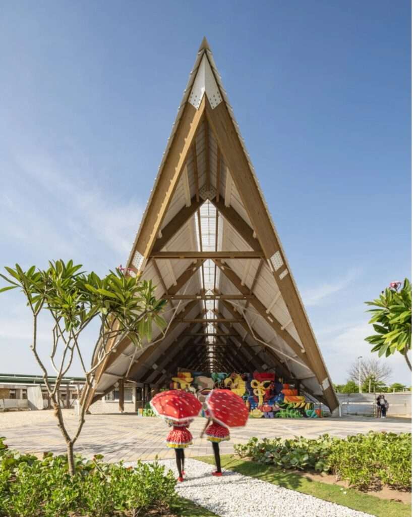 Symmetrical front view of the tall A-frame timber structure of the Vilas de San Pablo Cultural Center with pedestrians in the foreground.