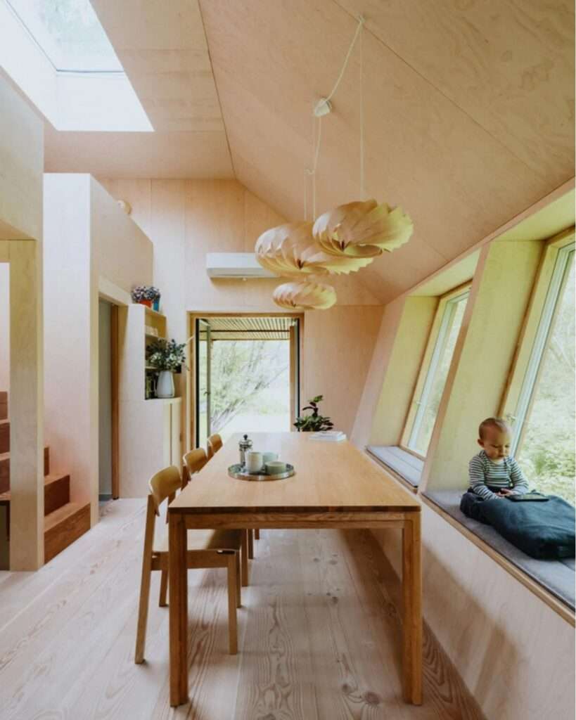 Bright double height plywood interior of EcoNeo cabin featuring a long dining table and child sitting by the window.