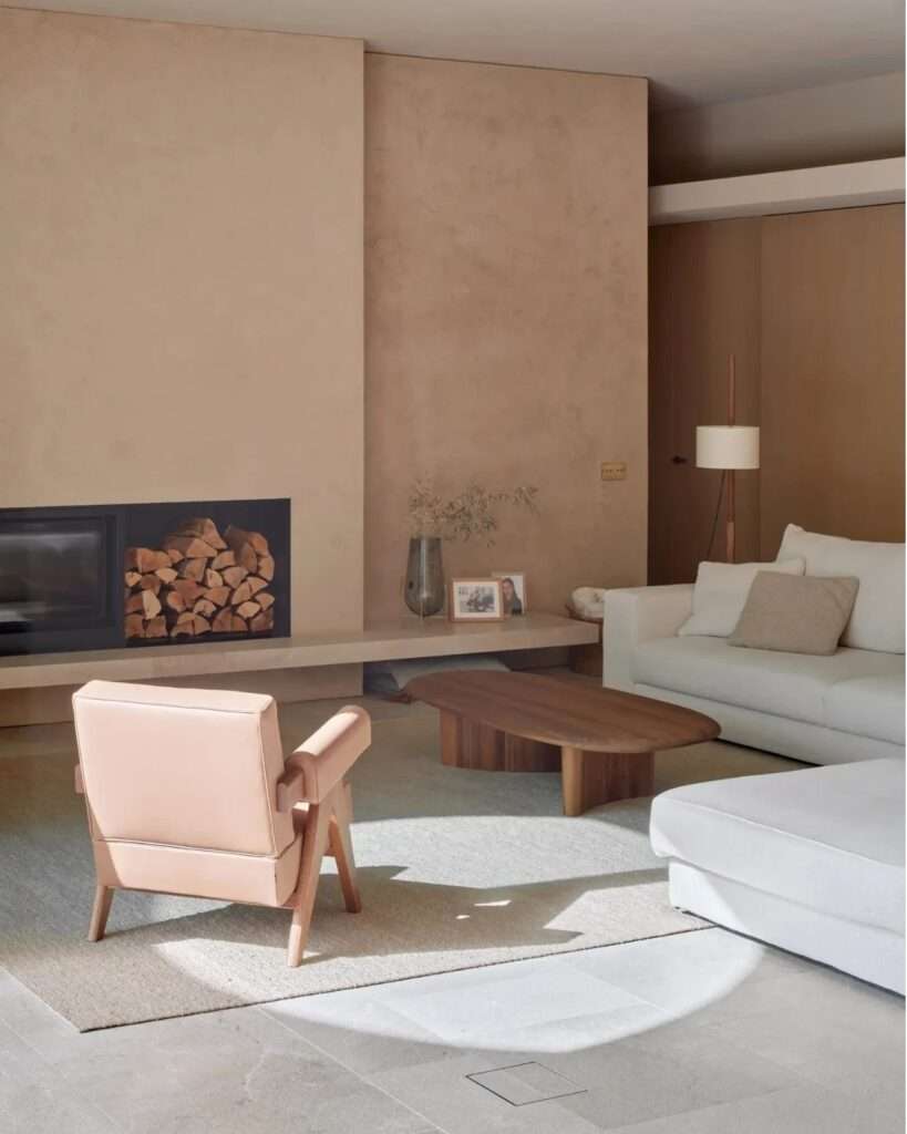 An interior view of a living area in Casa Óculo, showing a double fireplace, a white sofa, and a natural leather accent chair arranged under a beam of sunlight.