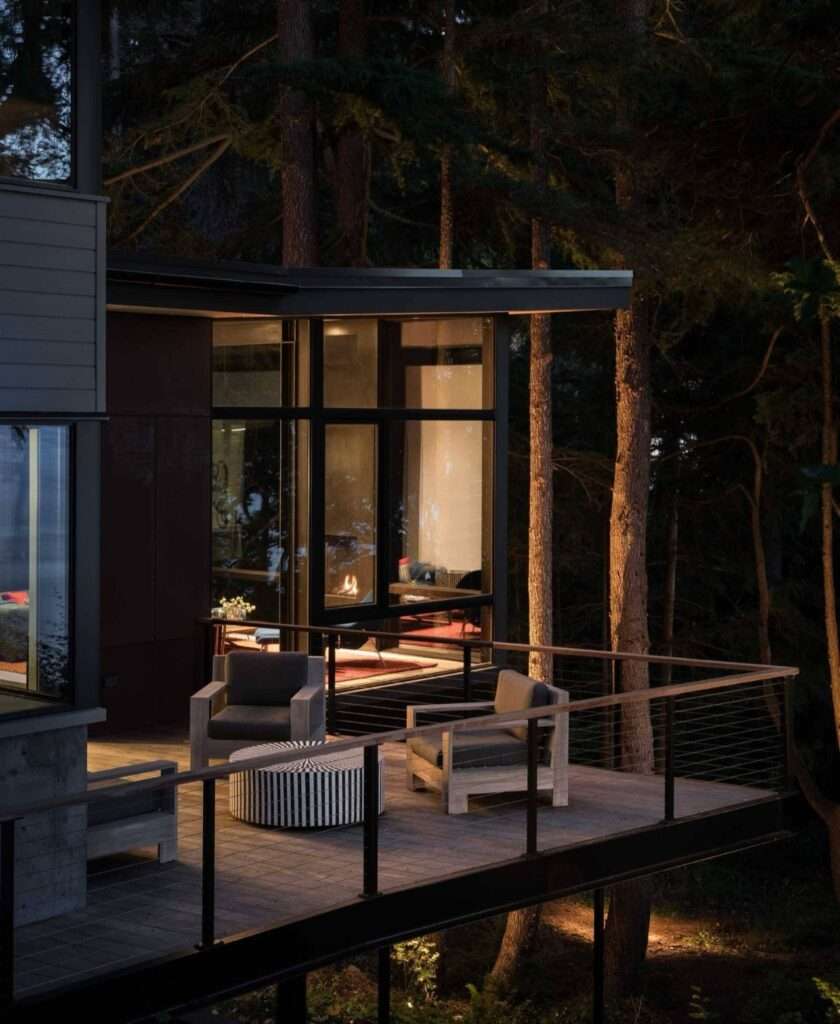 Cantilevered wooden deck with outdoor chairs lit up at night surrounded by tall pine trees.