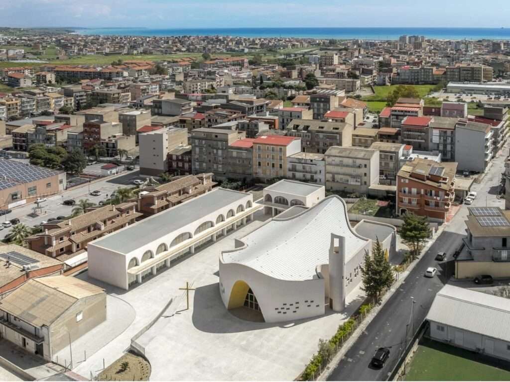 Aerial drone photograph of the Santa Barbara Parish Complex showing its linear layout, courtyard, church, and adjacent arched community center in Licata near the sea.