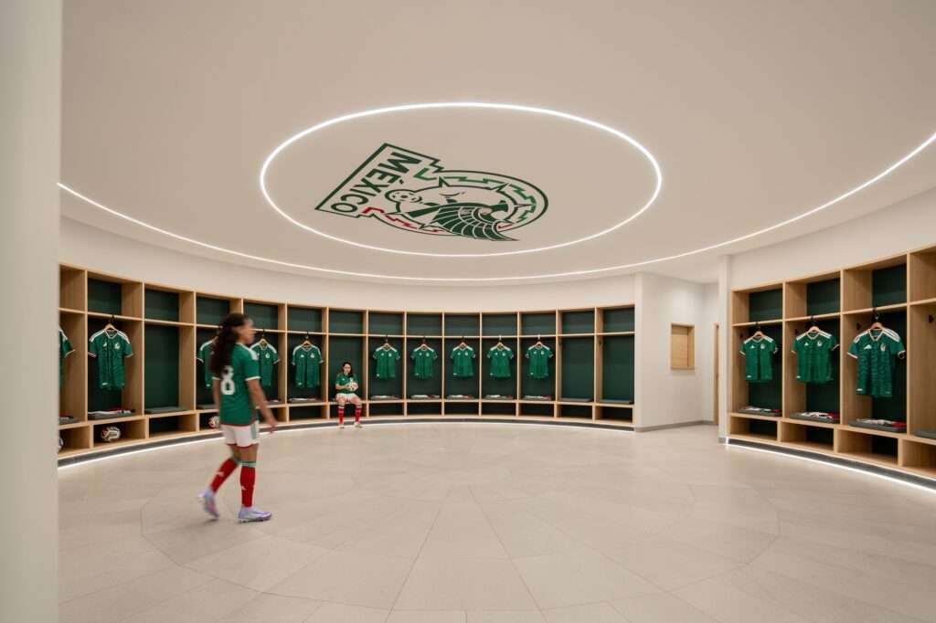 Circular locker room interior with wooden open lockers displaying green Mexico national football team jerseys beneath a recessed circular ring light and ceiling graphic.
