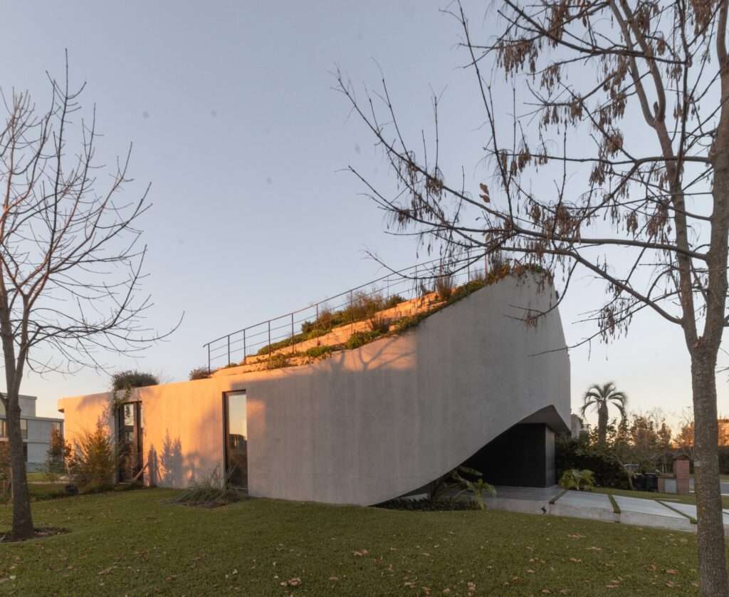 A low-angle architectural shot of a curved concrete residential building at golden hour, showcasing a sloped green ramp with step stairs and warm sunlight reflections.