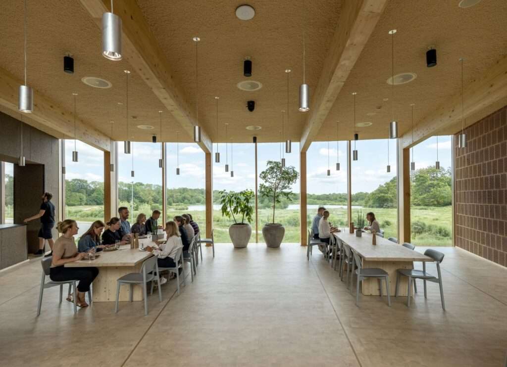 Employees dining at long wooden tables in the bright Dymak HQ cafeteria overlooking a green landscape.