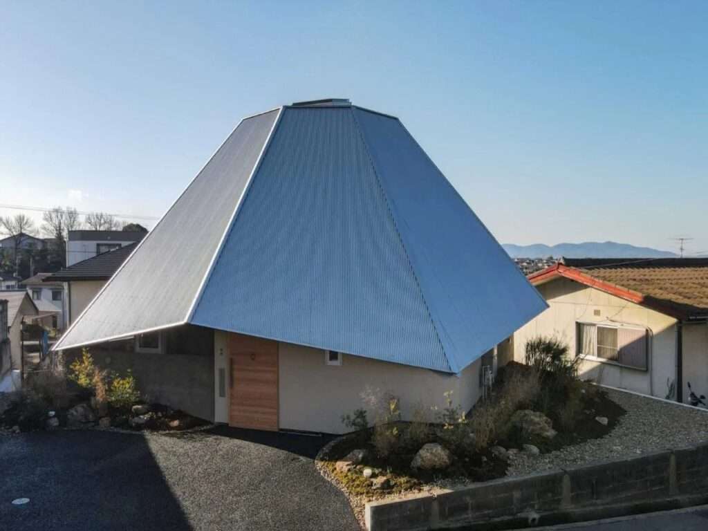 Exterior view of a modern house with a large, asymmetrical, multi-faceted metallic roof structure situated on a sloped residential plot.
