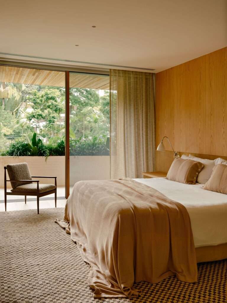 Cozy bedroom in Jacarezinho II House featuring a wooden wall headboard, neutral-toned bedding, a mid-century armchair, and a sliding glass door opening to a green balcony.