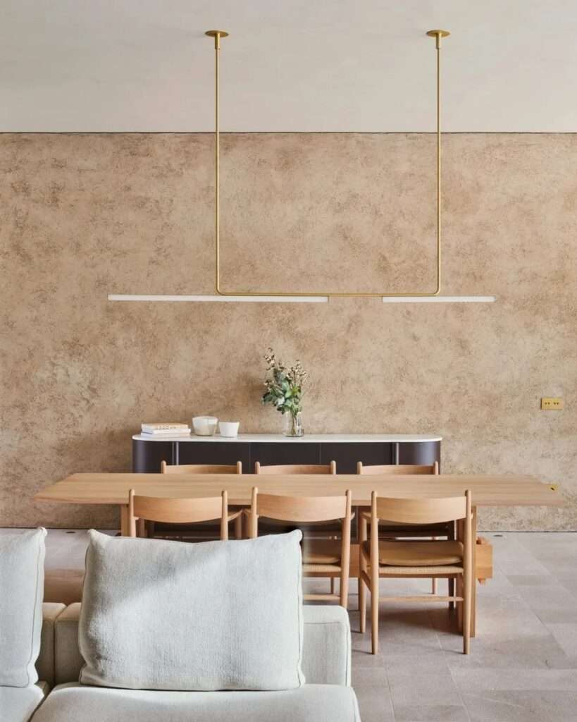A detail of a minimalist dining area in Casa Óculo, featuring a natural wood table, matching chairs, and a sleek brass pendant light against a textured wall.