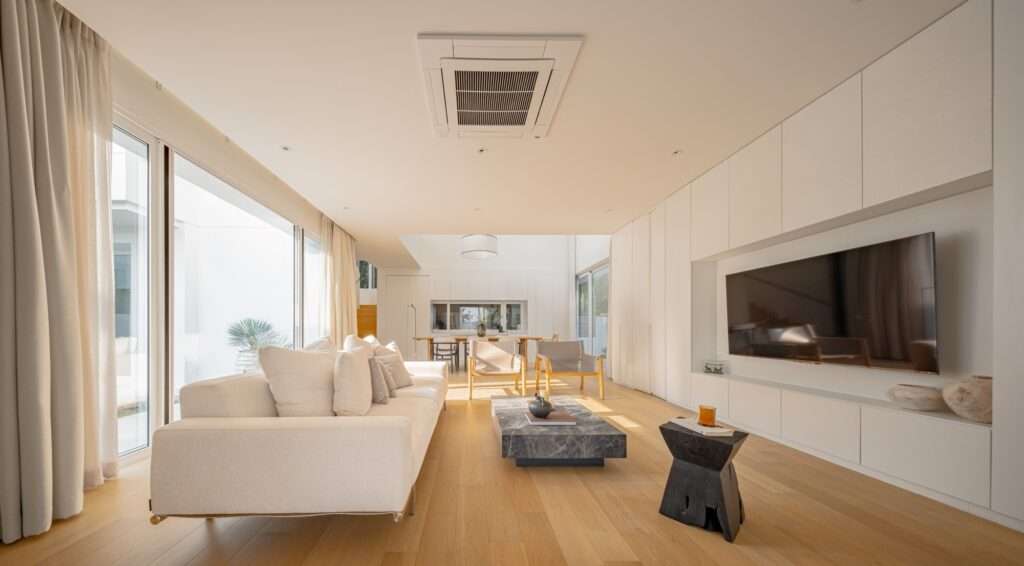 Minimalist open-plan living room interior of JNG House featuring a white sofa, warm wood flooring, integrated full-height storage panels, and large glass windows.