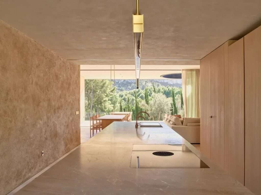 Interior view of an open-plan kitchen island with copper fixtures looking out through large glass openings toward lush Mediterranean pine hills.