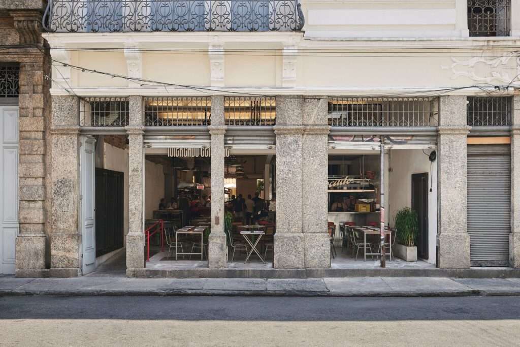 Close-up of the ground floor facade and main entrance of the Fábrica Bhering adaptive reuse in Rio de Janeiro, Brazil. Large, floor-to-ceiling windows with stone frames line the street, offering a glimpse inside to a restaurant area with tables and chairs. The floor has a raw, unfinished look. Photographed by Rafael Salim.