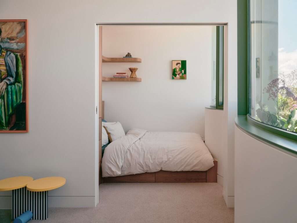 Minimalist bedroom alcove with a built-in timber bed frame, curved shelving, a pocket sliding door, and a view through a curved green glass window.