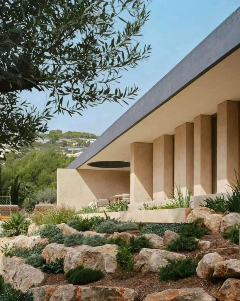 Exterior low-angle view of Casa Óculo showing a deep concrete roof overhang, vertical shade fins, and a terraced rock garden.