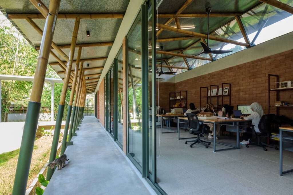 Active architectural studio with employees working at desks, framed by angled structural bamboo pillars along a covered concrete walkway and floor-to-ceiling glass doors.