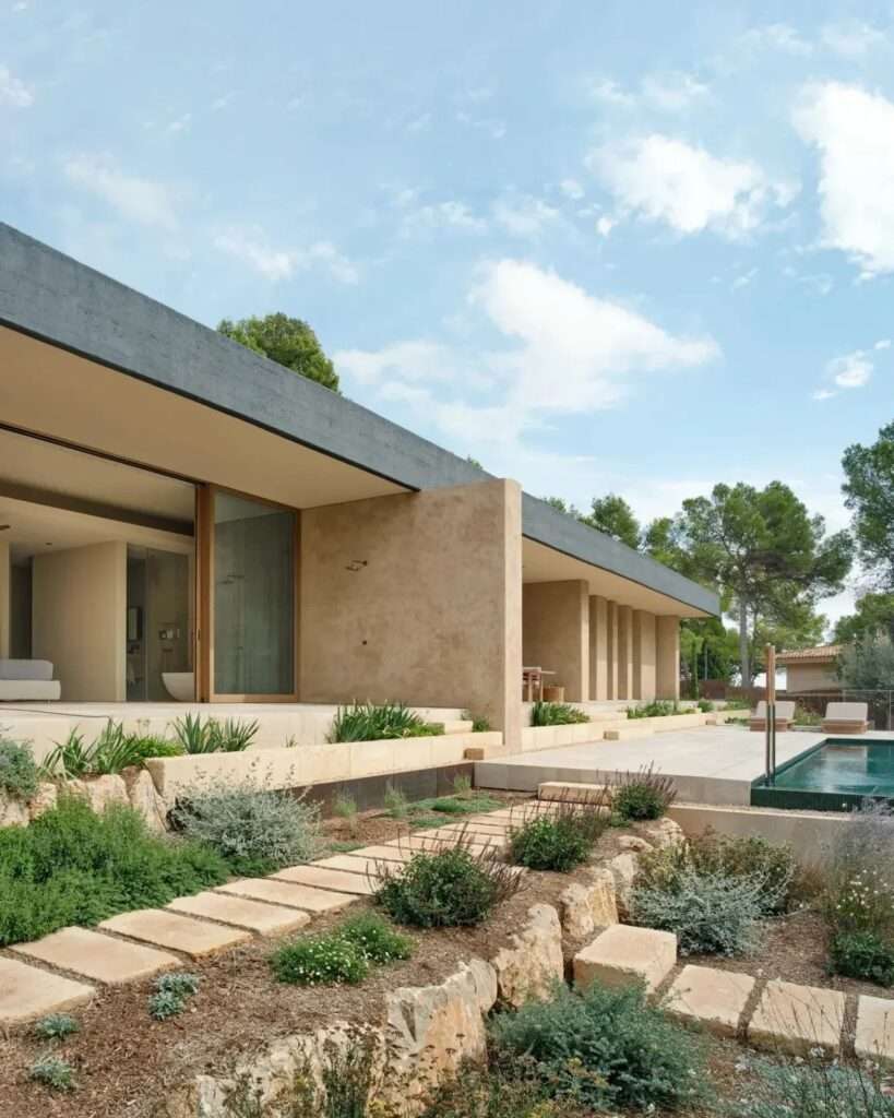 Exterior side view of Casa Óculo featuring stepped stone pathways, natural lime mortar walls, sliding glass doors, and a swimming pool deck.