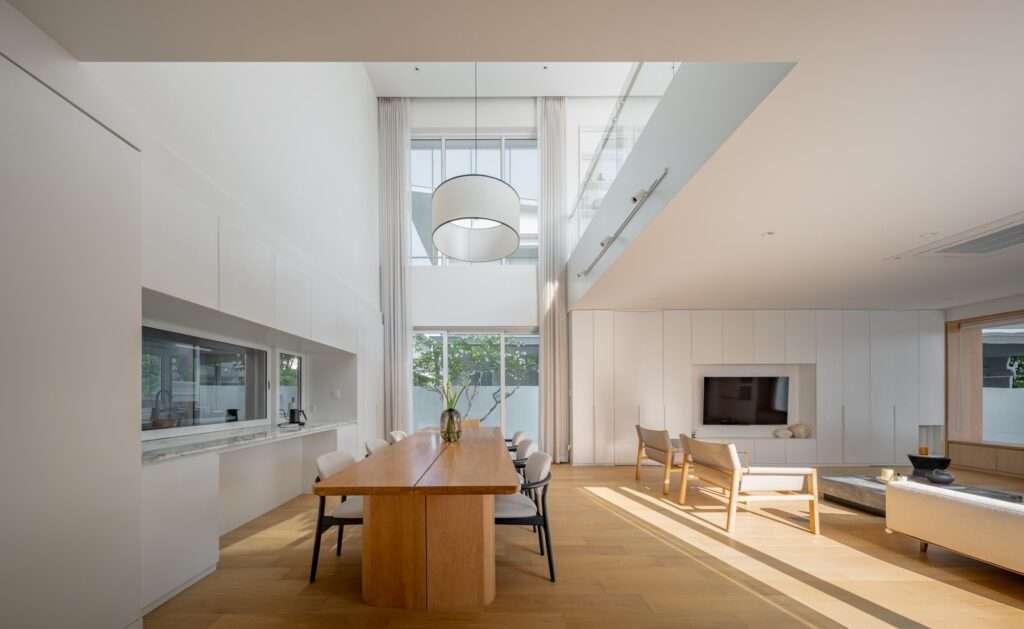 Double-height dining space at JNG House with a long wood table, modern minimalist kitchen counters, a large pendant light, and a second-floor mezzanine balcony.