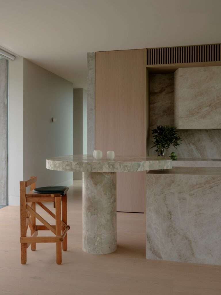 Modern kitchen featuring a light oak cabinetry wall and a custom stone counter island with a cylindrical leg and wooden bar stool.