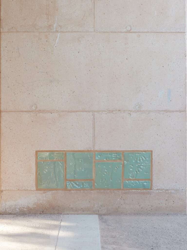 Close-up of handmade green-glazed ceramic tiles featuring children's handprints embedded into a bush-hammered concrete wall.