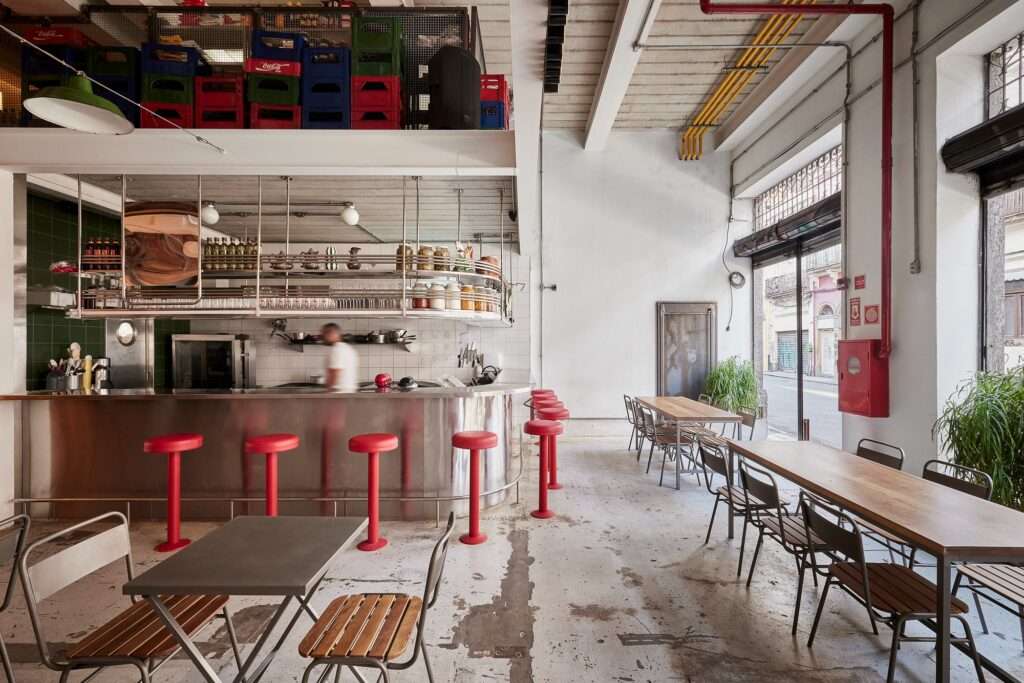 Interior of a restaurant area within the Fábrica Bhering adaptive reuse in Rio de Janeiro, Brazil. The space features a polished concrete floor with a distressed pattern, exposed white walls, and a large central bar made of stainless steel with overhead storage and red bar stools. Photographed by Rafael Salim.