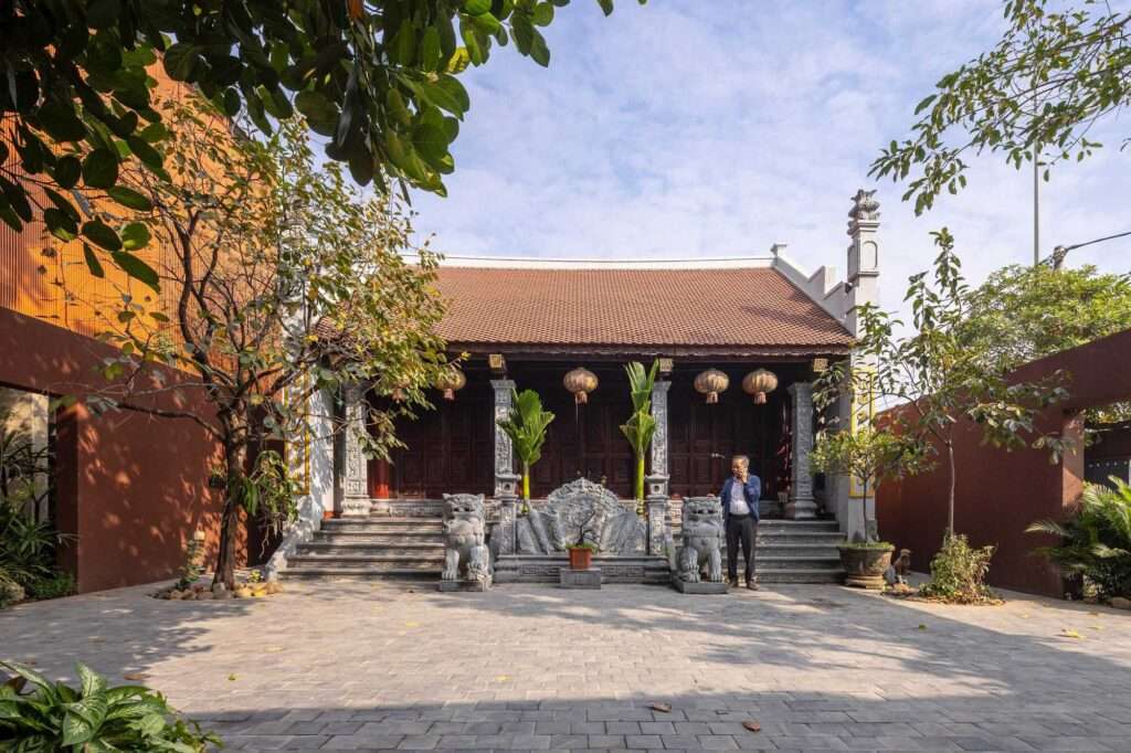 A man standing in the paved courtyard of NB01 House in front of the traditional family altar house with ornamental stone lanterns and carvings.