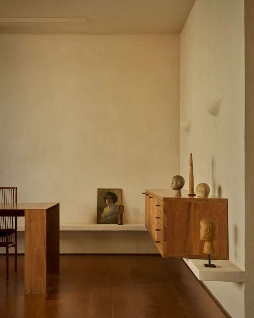 Dining and wall sideboard area featuring a floating wooden credenza on a low concrete ledge, adorned with small sculpted heads, next to a dining table and an oil painting resting against the wall.