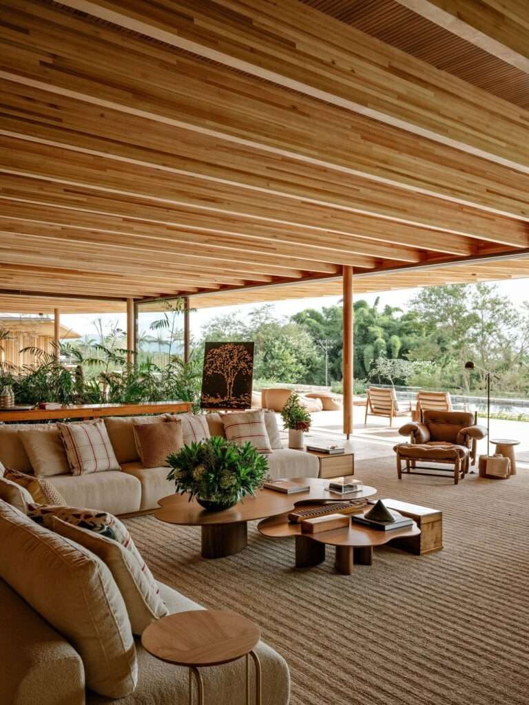 Interior design of AMS House living room featuring an exposed timber ceiling, neutral contemporary sofas, organic wooden coffee tables, and a leather armchair overlooking the forest.