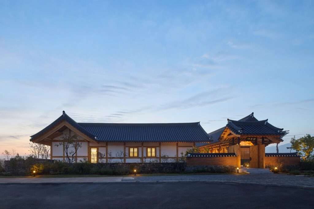 Wide exterior twilight view of the complete JK-AR timber pavilion complex and courtyard walls illuminated by subtle landscape lighting against a blue evening sky.