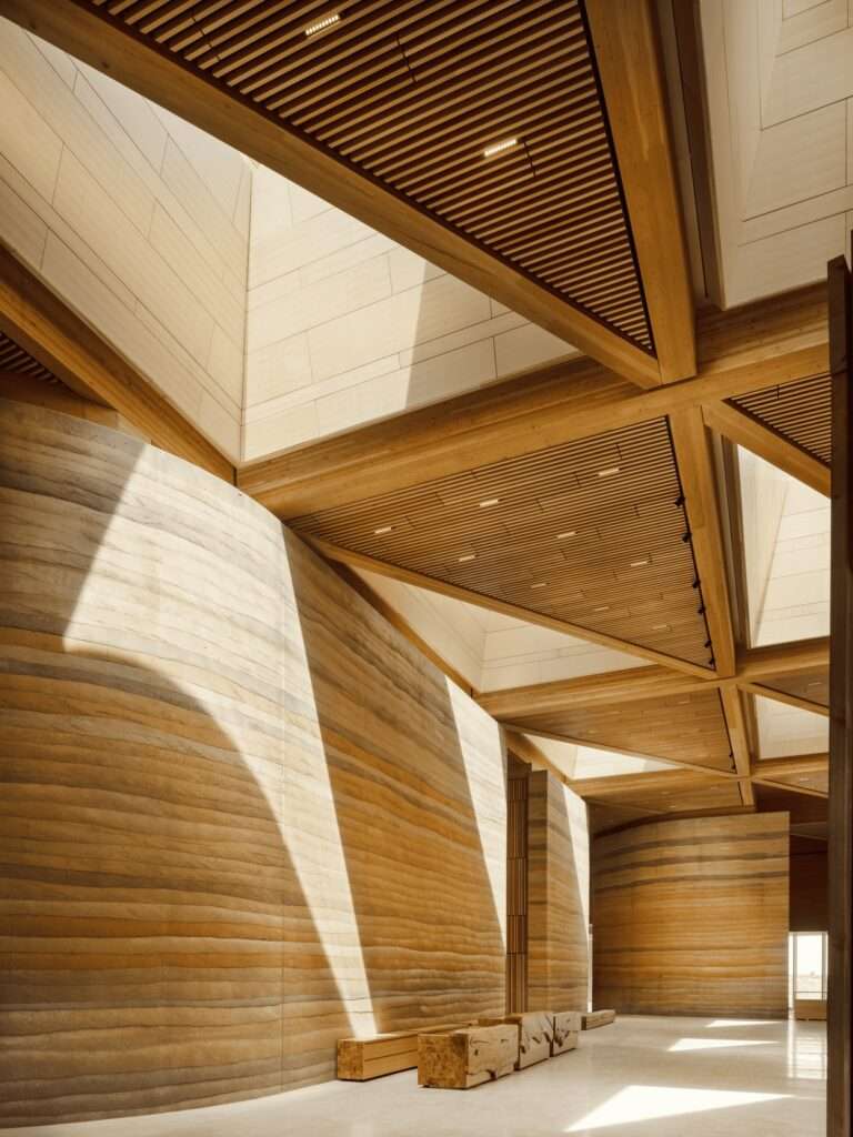An interior corridor featuring massive rammed earth walls with colorful geological layers and geometric timber ceilings.