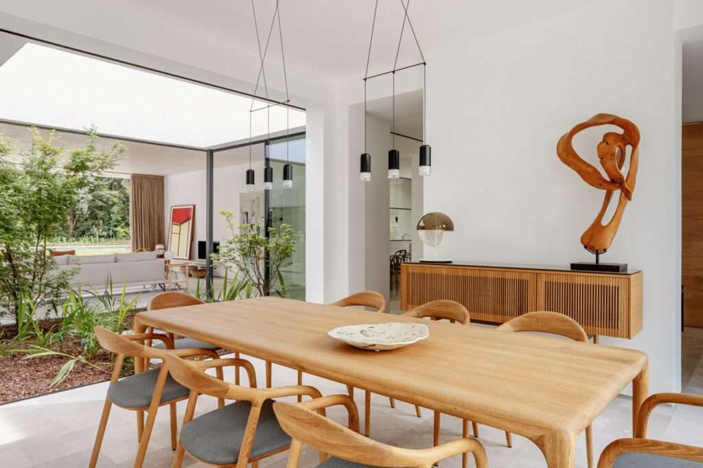 Interior dining space with a large light wood table and Scandinavian-style chairs, minimalist pendant lights, and a unique modern wooden sculpture on a sideboard, adjacent to the central courtyard.