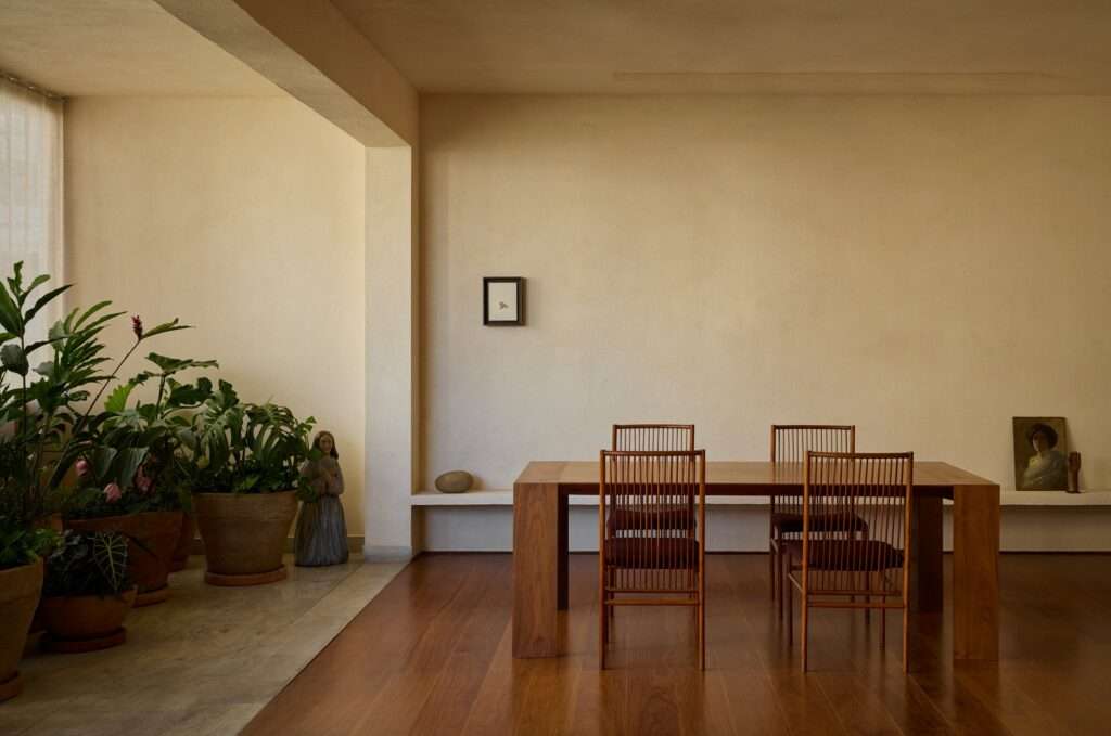 Minimalist wooden dining table with four slatted wooden chairs set on dark wood flooring, adjacent to an indoor garden with potted plants and a small decorative statue.