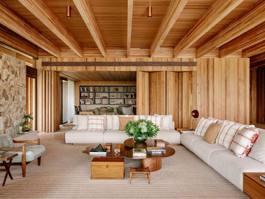 Spacious open-plan living room in AMS House showing light fabric sofas, organic coffee tables, a rustic stone wall accents, and warm wooden paneling.