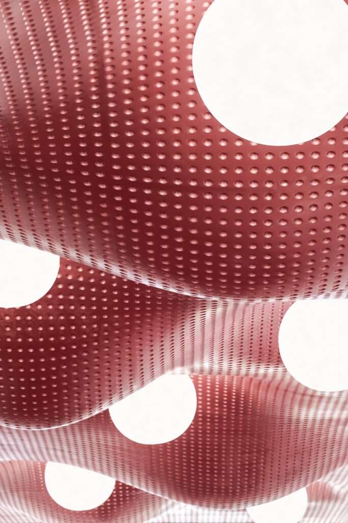 Abstract close-up of a red perforated three-dimensional wavy ceiling panel with circular cutouts for recessed lighting fixtures at Wuxi MixC Mall.
