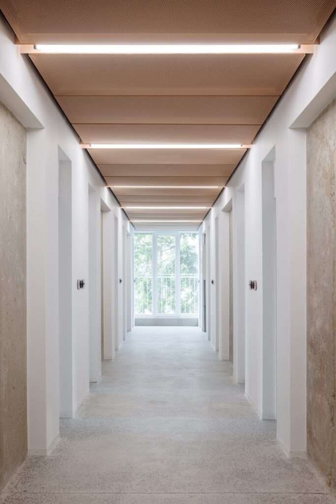 Long, brightly lit minimalist apartment corridor with white walls, exposed raw concrete edges, and a perforated copper-tone mesh ceiling.