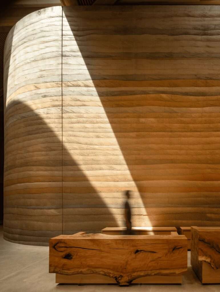 Close-up of the rammed earth wall texture showing distinct horizontal geological layers with a blurred figure walking past.