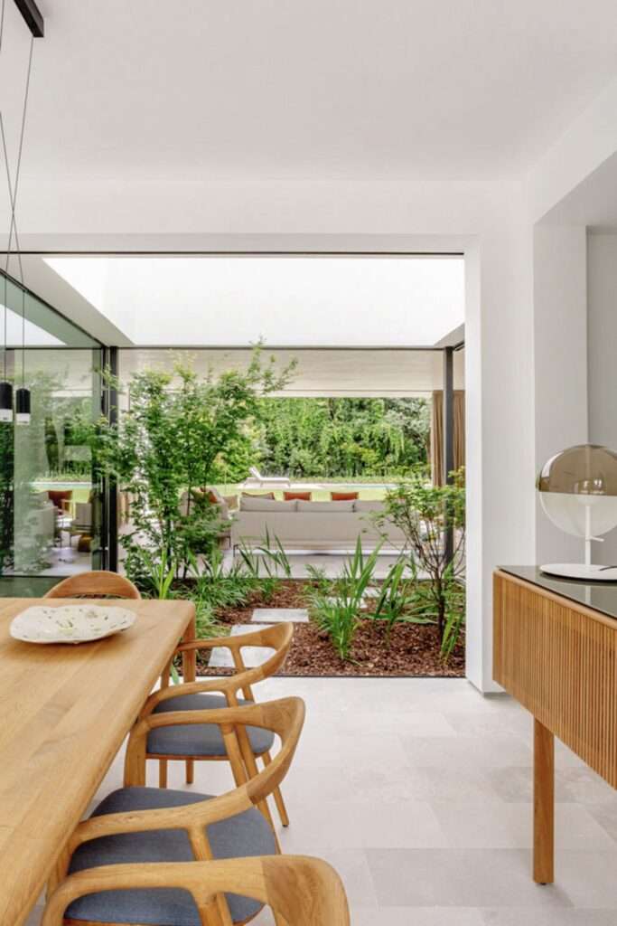 A view across the wooden table and chairs of the dining area, through the central glass courtyard filled with trees, towards the large outdoor lounge sofa and the rear garden and pool.