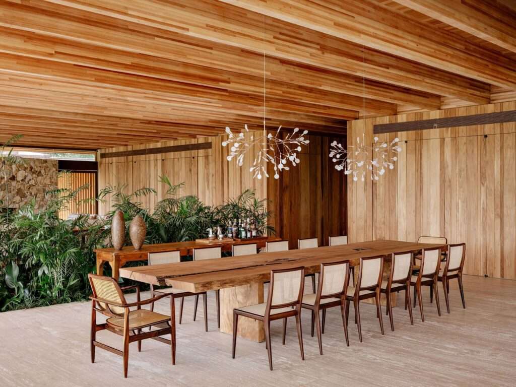 Modern dining room inside AMS House featuring a long live-edge solid wood dining table, mid-century modern cane chairs, delicate white chandeliers, and indoor tropical plants.