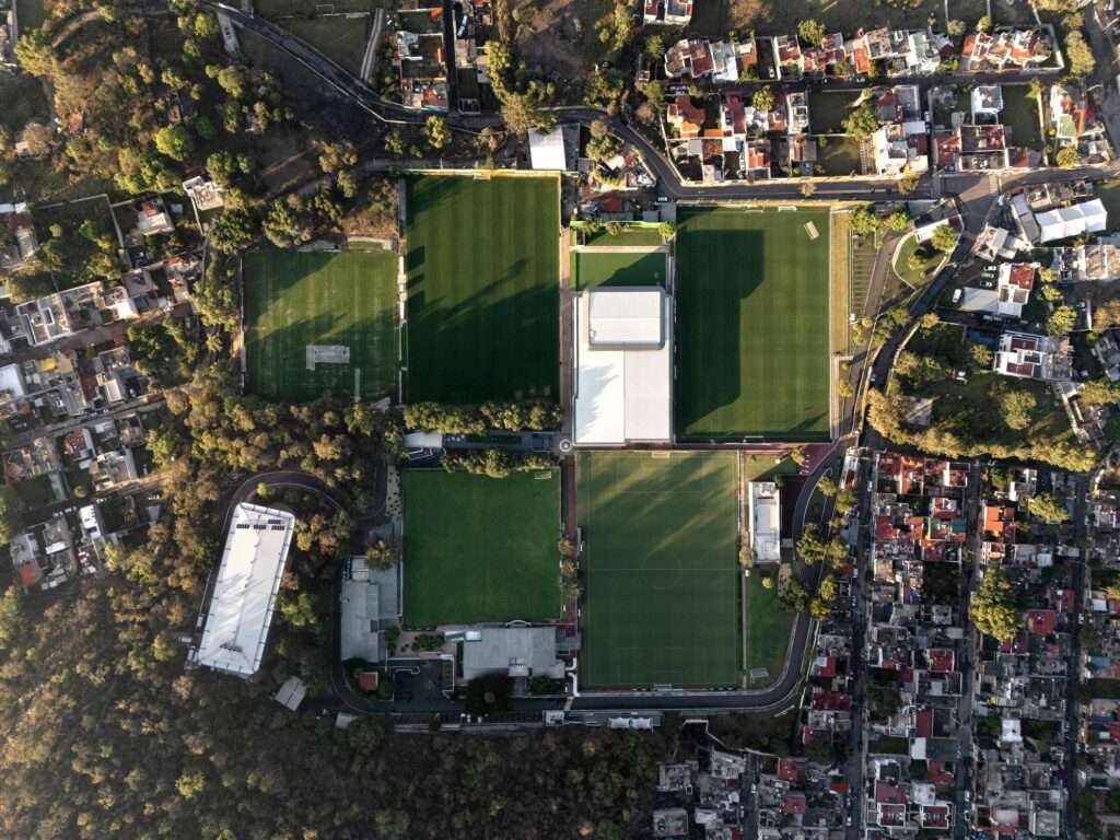 Top-down orthographic aerial view of the entire high-performance center master plan, showing multiple green soccer pitches, low-rise structures, and the dense urban-nature border.