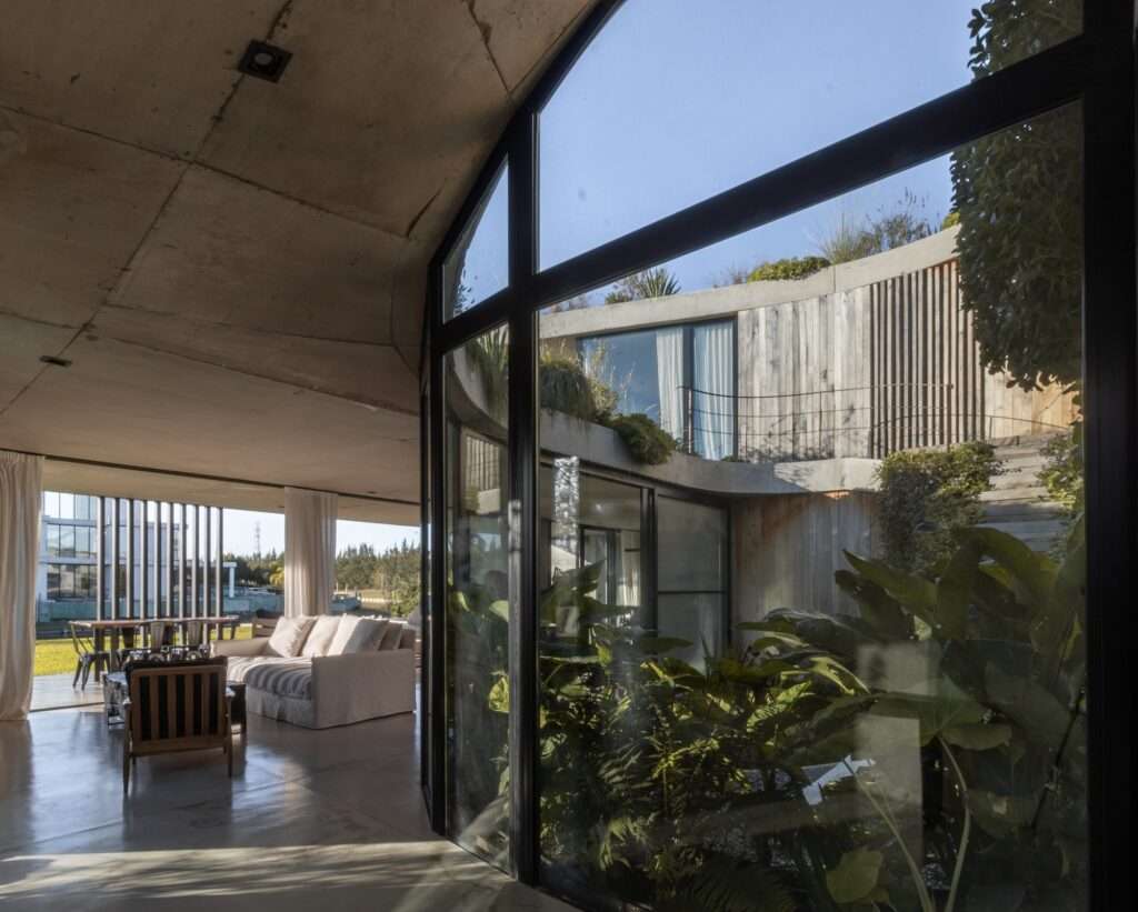 A dramatic low-angle interior shot looking past a structural glass frame into a multi-level open courtyard with timber stairs and tropical plants.