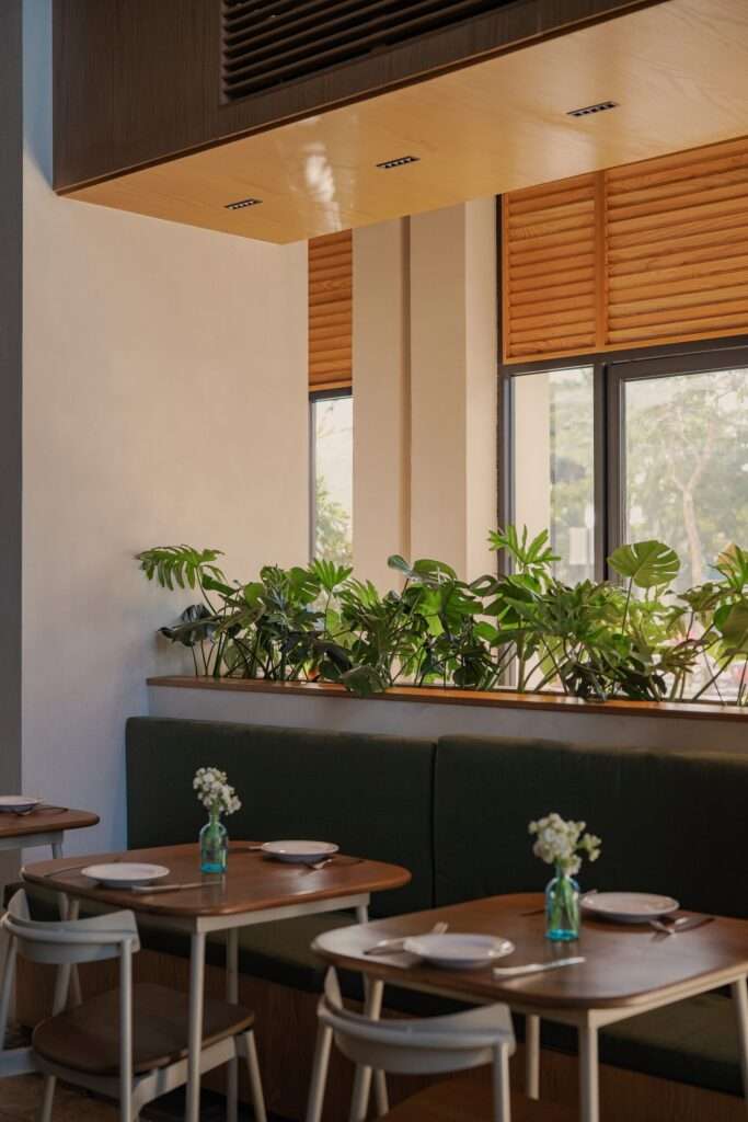 Intimate banquette seating with dark green upholstered cushions, oak tables, small floral vases, and window planters.