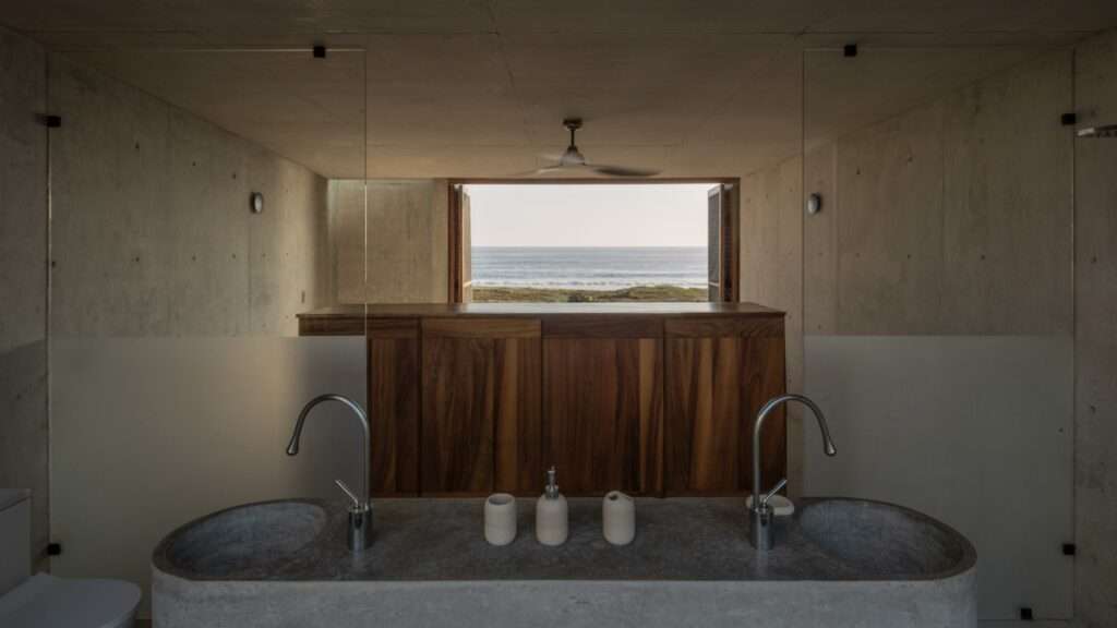 Minimalist concrete bathroom with dual vessel sinks, glass partitions, and a direct window looking out to the ocean.
