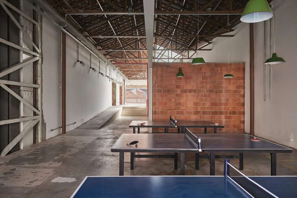 Interior view of a large, open space within the Fábrica Bhering adaptive reuse in Rio de Janeiro, Brazil. The space has high ceilings with exposed wooden trusses, whitewashed walls, and a ramp and stairs leading to an upper level. Three ping-pong tables are set up on a raw concrete floor. Photographed by Rafael Salim.