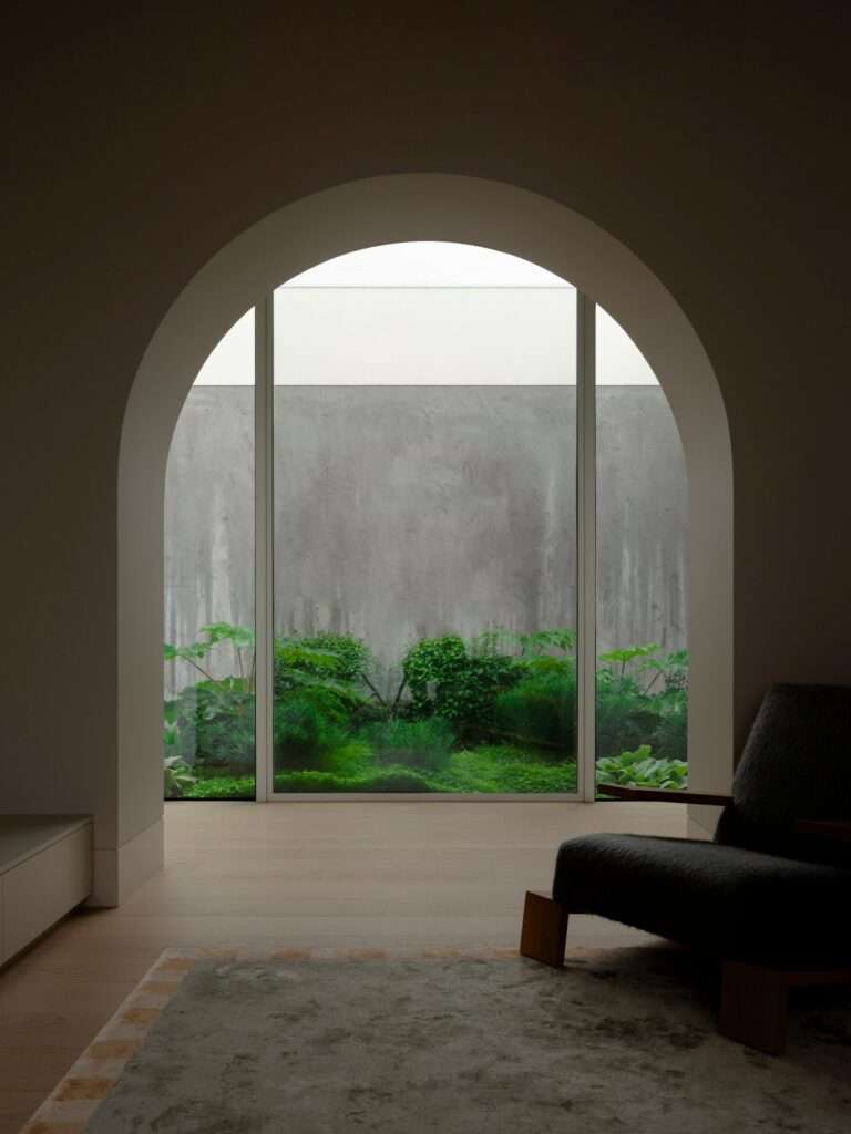 Interior lounge view framed by an arched architectural opening facing a full-height glass window looking out onto a lush green courtyard wall.